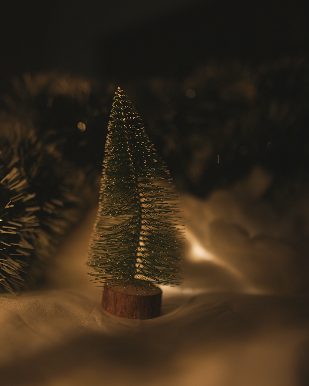 Small artificial Christmas tree on a wooden base, with a snowy background and warm lighting.