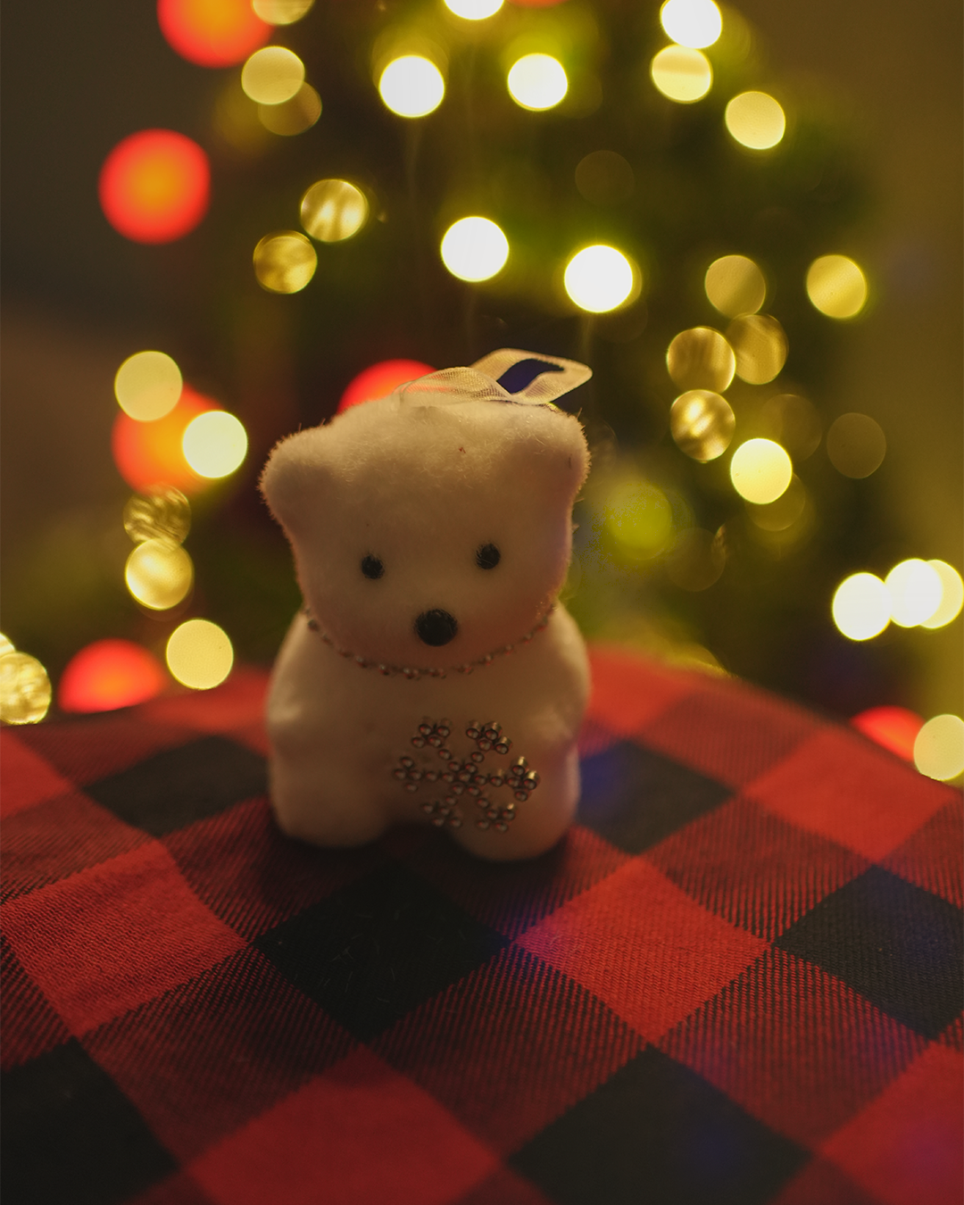 White plush Christmas bear ornament with a snowflake and gold accents, against a festive bokeh background.