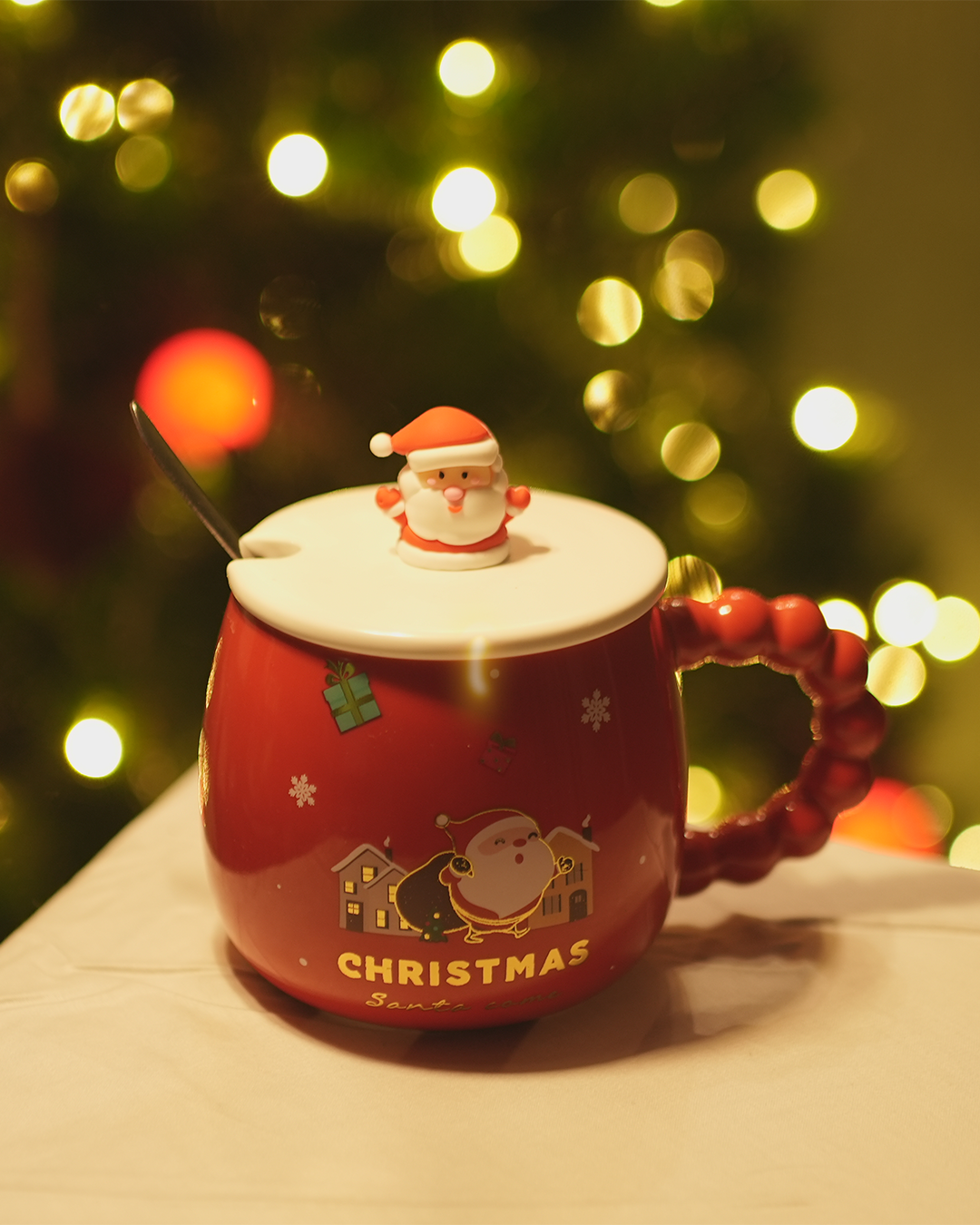 Red ceramic Christmas mug with a Santa Claus lid, spoon, and festive design, set against a backdrop of twinkling Christmas lights.